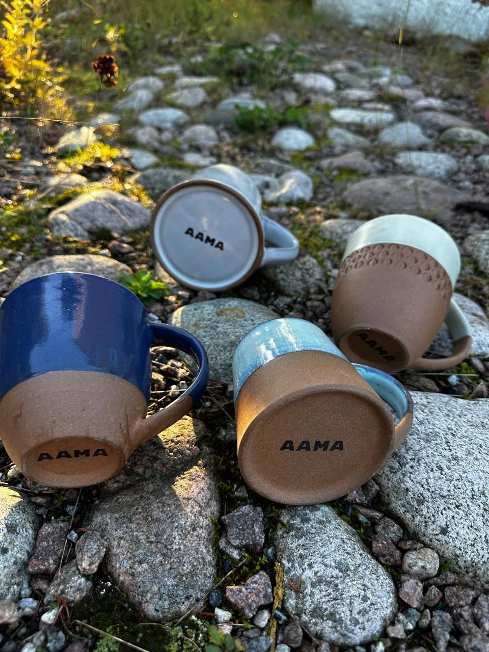 Handcrafted Aama stoneware mugs with blue and natural clay finishes, resting on stones outdoors